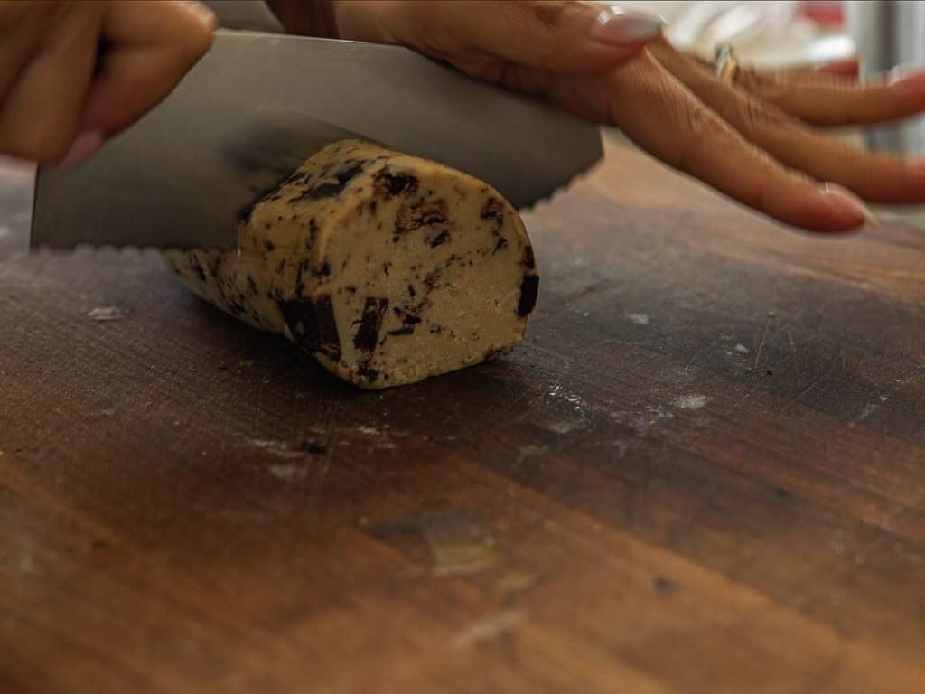 A person slices a log of vegan chocolate chip cookie dough with a large knife on a wooden cutting board.